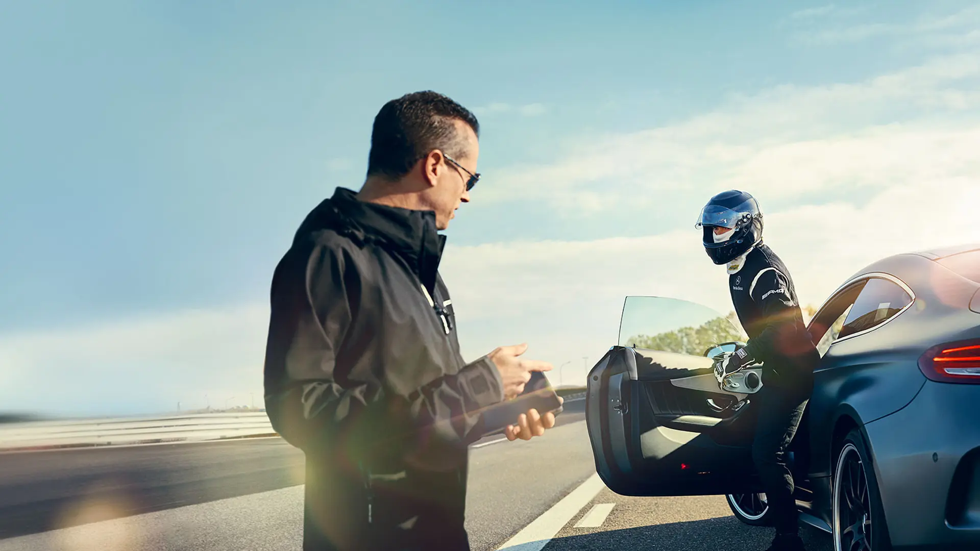 A man holding a phone greets a helmed driver leaving an AMG vehicle